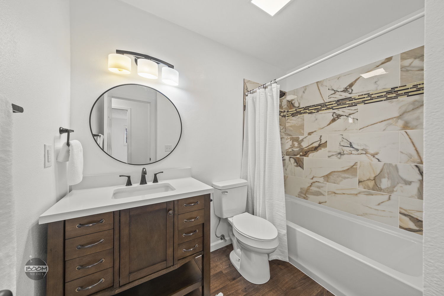 Guest bathroom with a wood vanity, round mirror and marble-look tub surround